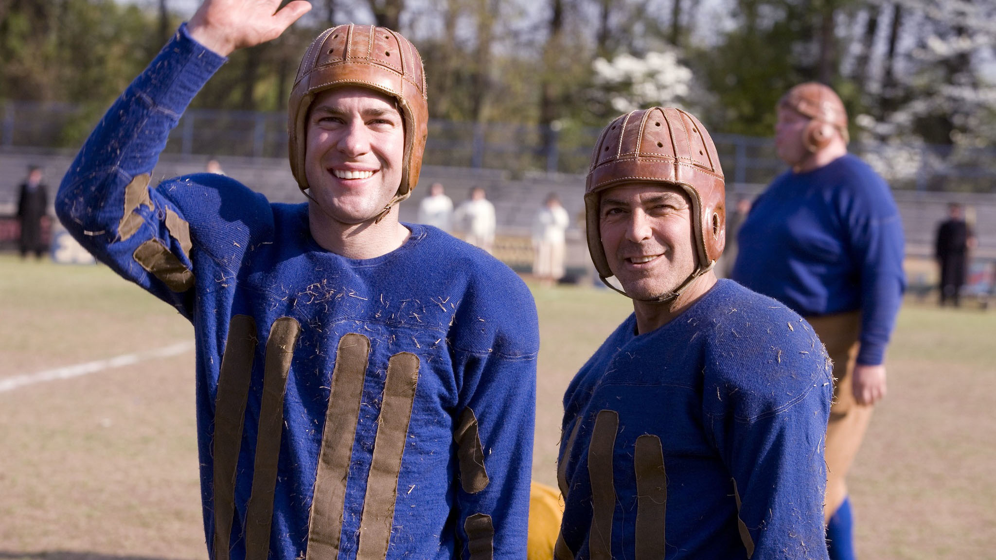 john krasinki and george clooney stand outside in blue football jerseys and brown leather helmets in a scene from leatherheads