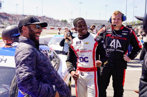NASCAR drivers Bubba Wallace and Rajah Caruth laugh while standing near cars on a race track